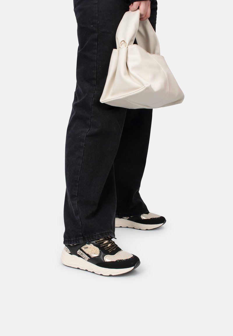 Jeans noirs, sac à main crème et baskets noir et blanc avec des panneaux en mesh et lisses, des accents dorés et des détails texturés sur le dessus.