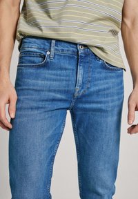 Blå denimjeans med smal passform, som har en klassisk femficksdesign, sydda sömmar och subtil blekning. Bärs med en randig, grön topp.