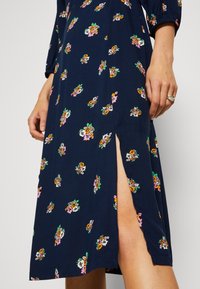Robe bleu marine avec de petits motifs floraux roses, orange et blancs, dotée d'une fente latérale dévoilant une partie de la jambe et deux mains près de la taille.