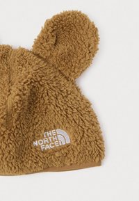 Barna, bolyhos plüss babasapka, medvés fülekkel, a fehér háttér előtt fehér hímzett The North Face logóval.