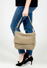 Vaalea beige käsilaukku, jossa on teksturoitu käärmeennahkakuosi, varustettuna läpän sulkemisella ja säädettävällä hihnalla. Laukkua pitelee henkilö, jolla on päällään sinisiä farkkuja.