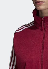 Bordeauxrote Zip-Jacke mit hohem Kragen und weißen Drei-Streifen-Akzenten über den Schultern. Hergestellt aus strukturiertem Stoff.