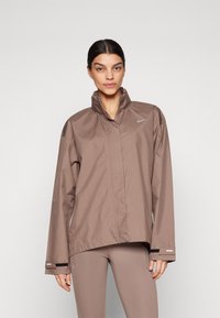 Smeđi Nike jakna s visokim ovratnikom, dugim rukavima i reflektirajućim detaljima. Izrađena od laganog materijala, s opuštenim krojem i jednostavnim dizajnom.
