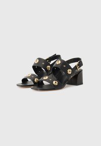 Sandali neri in pelle con dettagli floreali dorati, punta aperta e tacco largo. Presentano una cinghia regolabile sul retro e un design traforato.