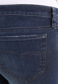 Mörkblå denimjeans med en bakficka som har ett V-format söm mönster. Tyget har en slät yta och en lätt blekning.