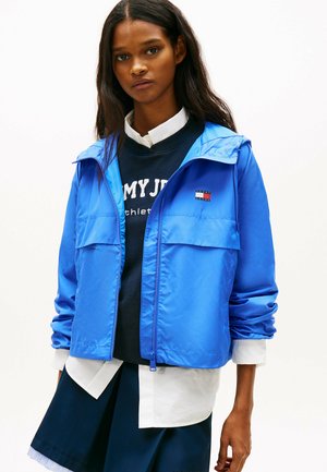 Junge Frau mit langem dunklem Haar, die eine leuchtend blaue Jacke mit Reißverschluss über einem marineblauen Sweatshirt und einem weißen Hemd trägt, vor einem schlichten hellen Hintergrund stehend.