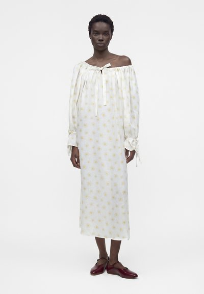 Γυναίκα με λευκό φόρεμα μέσης με έξω από τον ώμο, κίτρινα λουλουδάτα σχέδια, μακριά μανίκια και μπορντό επίπεδα παπούτσια.