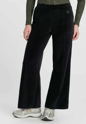 Persona che indossa pantaloni neri a gamba larga, scarpe da ginnastica argento e un top verde a maniche lunghe con motivo leopardato.