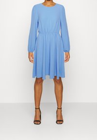 Hellblaues langärmliges Kleid mit rundem Ausschnitt, elastischem Taillenbund und fließendem Rock. Getragen mit schwarzen Riemchen-Heels. Glatte Textur.
