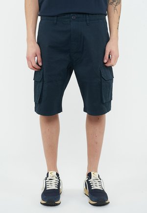 Uomo che indossa pantaloncini cargo blu navy con tasche laterali con bottoni e sneakers blu navy con lacci bianchi, in piedi contro uno sfondo semplice.