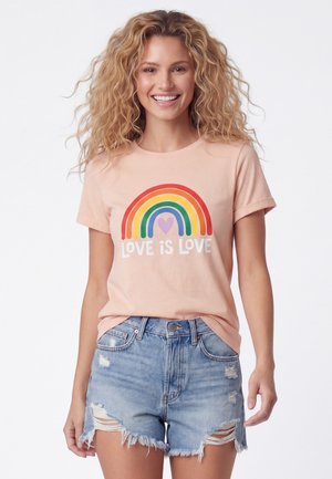 Jonge vrouw met krullend haar draagt een perzik T-shirt met regenboog en de tekst ""Love is Love"", gecombineerd met versleten spijkershort.