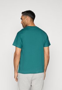 T-shirt verde-água de manga curta exibida de costas, com decote redondo, bainha reta e textura de algodão suave. Combinada com calças cinzas.