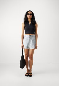 Svart glidelås uten ermer topp, lyseblå denimshorts, svart veske og svarte plattform sandaler, med krøllete hår og solbriller.