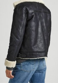 Veste en cuir synthétique noir avec col et poignets en shearling, présentant un design élégant et une finition texturée, associée à un jean bleu.
