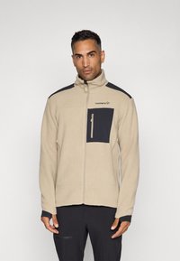 Beige fleecejakke med sorte detaljer, høy krage og glidelåslomme. Har en teksturert overflate og en figursydd design.