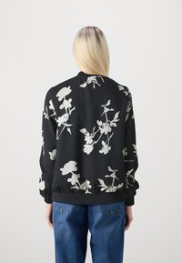 Person med långt blont hår som bär en svart bomberjacka med vit blommönstring och blå jeans, vänd bort från kameran mot en enfärgad bakgrund.