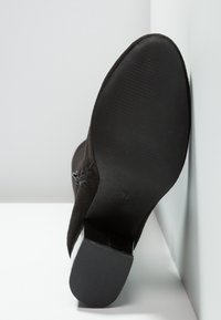 Bottine en daim noir avec un talon incurvé, une semelle noire lisse et une semelle extérieure texturée. Dispose d'une fermeture éclair latérale pour un enfilage facile.