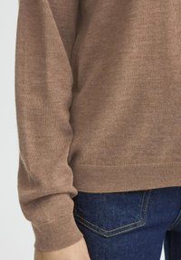 Jasnobrązowy sweter z dzianiny o luźnym kroju, z prążkowanymi mankietami i dołem, zestawiony z ciemnoniebieskimi dżinsami z detalami w szyciu.