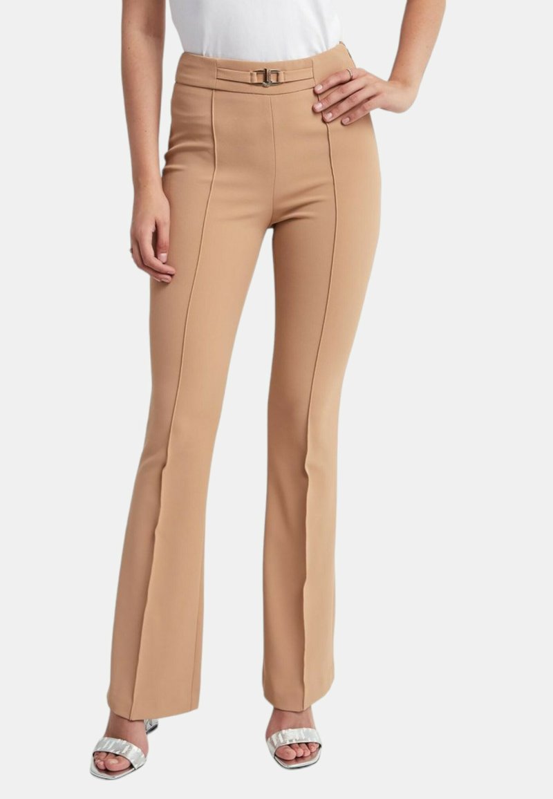Femme portant un pantalon évasé taille haute couleur camel avec des coutures frontales et une petite boucle de ceinture assortie, associé à des talons blancs ouverts à l'avant.