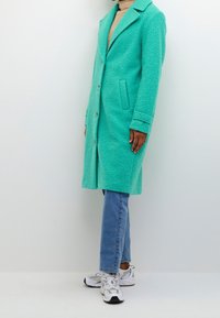 Cappotto turchese texture con colletto a punte, chiusura a bottoni e tasche laterali, indossato sopra jeans blu e sneakers bianche.