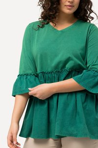 Grüne Baumwollbluse mit V-Ausschnitt, Rüschensäumen und gerafftem Taillenbund. Der Stoff hat eine weiche Textur und einen lässigen Schnitt.