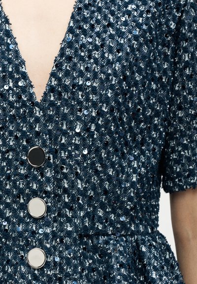 Gros plan sur une robe en tissu texturé bleu foncé avec des paillettes scintillantes, de grands boutons ronds argentés, des manches courtes et un décolleté en V.