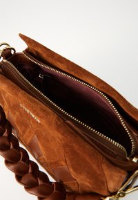 Brown suede handbag with a braided leather strap, zipped top, and a smooth inner lining. Features a gold zipper and textured body design.