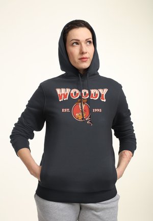 Kvinde iført en sort hættetrøje med teksten "WOODY EST. 1995" og en tegneserie-cowboy, med hænderne i lommerne, kigger op mod højre.