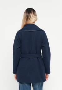 Femme aux cheveux blonds longs portant un manteau ceinturé bleu marine et un jean, de dos, devant un fond blanc uni.