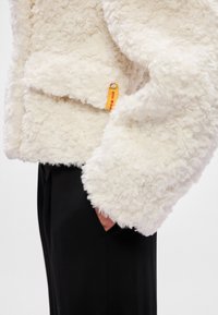 Λευκό ψεύτικο γούνινο μπουφάν με μαλακή, υφασμάτινη επιφάνεια, που διαθέτει ορατή πορτοκαλί ετικέτα. Μαύρο παντελόνι μερικώς ορατό κάτω από το μπουφάν.