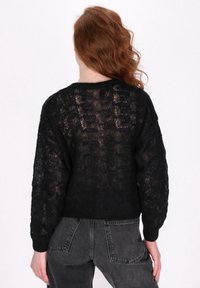 Maglione nero a maglia caratterizzato da un design loose e testurizzato, con una schiena semi-trasparente, maniche lunghe e polsini a costine. Abbinato a jeans grigi.