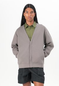 Grå sweatshirt med V-krage, långa ärmar och sidofickor, tillsammans med en grön skjorta med knappar och svarta shorts. Slät textur.