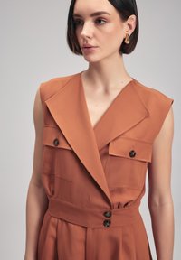 Rustfarbenes ärmelloses Kleid mit tiefem V-Ausschnitt, zwei Brusttaschen und tailliertem Schnitt. Verfügt über große Knöpfe und einen strukturierten Revers.