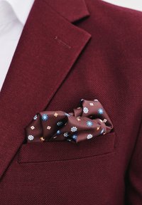 Bordowy, strukturalny żakiet z białą koszulą i brązowym, wzorzystym chusteczką w kieszeni, na którym znajdują się niebieskie i pomarańczowe kwiaty oraz geometryczne kształty.