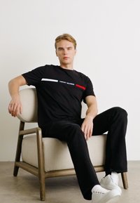 Schwarzes T-Shirt mit horizontalen weißen und roten Streifen, mit dem Text "TOMMY HILFIGER". Modell sitzt auf einem hellen Stuhl, trägt schwarze Jeans und weiße Sneakers.