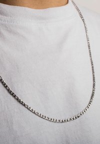 Strieborný náhrdelník z reťazí so prepojenými článkami, nosený cez jednoduché biele tričko. Dizajn je elegantný s hladkým povrchom.