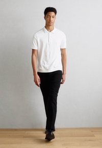 White polo shirt with a classic collar, paired with fitted black trousers and dark shoes, shown against a light background.