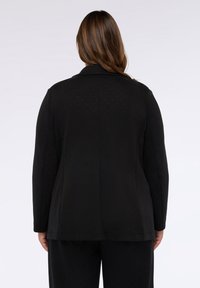Blazer nero con design strutturato, caratterizzato da una texture a pois, colletto a revers e maniche lunghe. La vista posteriore mostra una vestibilità su misura e linee pulite.