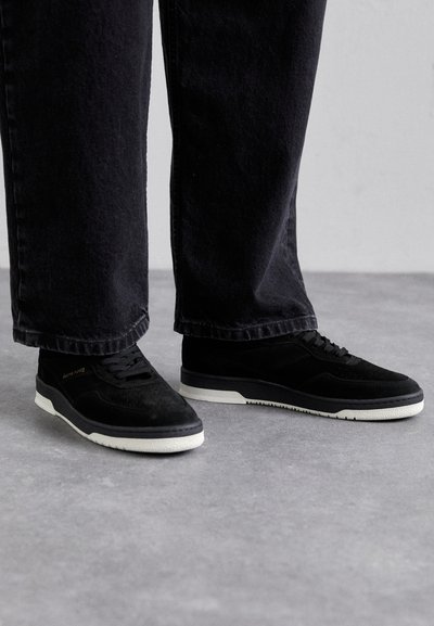 Schwarze Wildledersneaker mit einer weißen Gummi-Sohle, ausgestattet mit perforierten Details, schwarzen Schnürsenkeln und einem minimalistischem Design. Dunkle Jeans sind teilweise sichtbar.
