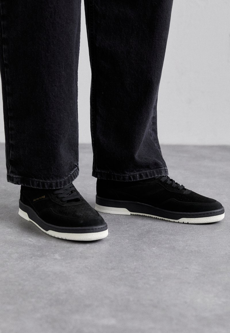 Black suede sneakers with a white rubber sole, featuring perforated detailing, black laces, and a minimalistic design. Dark jeans partially visible.