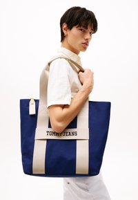 Blå canvas shoppingväska med beige remmar, som har en framträdande "TOMMY JEANS"-logotyp. Strukturerad design och slitstarkt material.