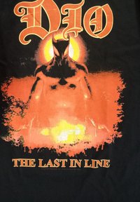 Černé tričko s oranžovou a žlutou grafikou svalnaté postavy na ohnivém pozadí, s nápisy "DIO" a "THE LAST IN LINE".