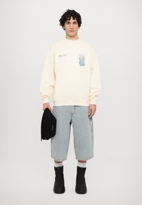 Cremefarbener Sweatshirt mit blauer Grafik, oversize Schnitt, kombiniert mit hellen Jeansshorts, schwarzer Tasche und schwarzen Ankle-Boots.