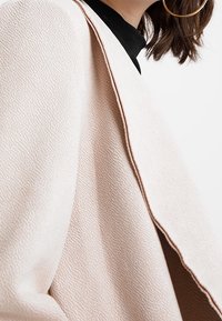 Donna che indossa un cappotto beige chiaro e strutturato sopra un top nero, con un grande orecchino a cerchio dorato e capelli castani lunghi fino alle spalle.