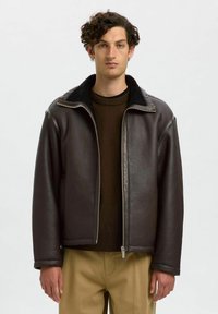 Brun läderjacka med dragkedja framtill, en krage i syntetisk päls och en avslappnad passform. Bärs över en mörk tröja, med beiga byxor synliga nedanför.