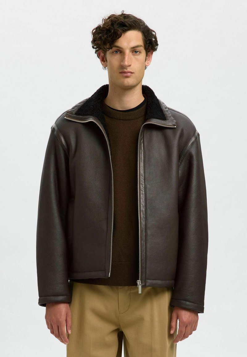 Brun läderjacka med dragkedja framtill, en krage i syntetisk päls och en avslappnad passform. Bärs över en mörk tröja, med beiga byxor synliga nedanför.