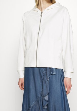 Femme portant un sweat à capuche blanc zippé avec un anneau et une jupe en jean bleue avec des détails à volants.
