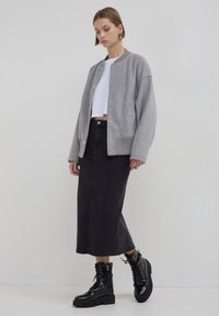 Frau in grauem oversized Blazer, weißem Cropped-Top, schwarzem Midikleid und schwarzen Schnürstiefeln, die vor einem schlichten Hintergrund steht.