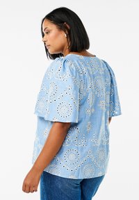 Blusa in cotone azzurro chiaro con maniche corte a palloncino, caratterizzata da ricami floreali bianchi e dettagli a intaglio, scollatura rotonda e vestibilità rilassata.