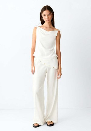 Femme debout portant un haut blanc sans manches avec des bretelles nouées aux épaules, un pantalon ample assorti et des sandales noires sur fond blanc.